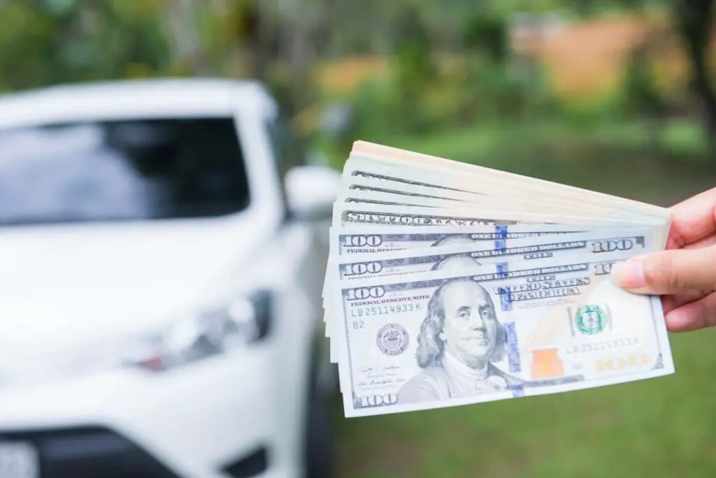 Hand holding 100-dollar bills in front of a white car
